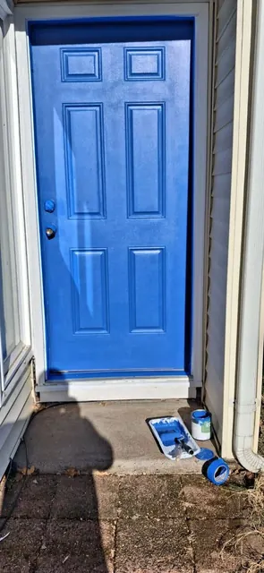 A paneled entry door has a bright blue finish, with masking tape and painting supplies still around the threshold.