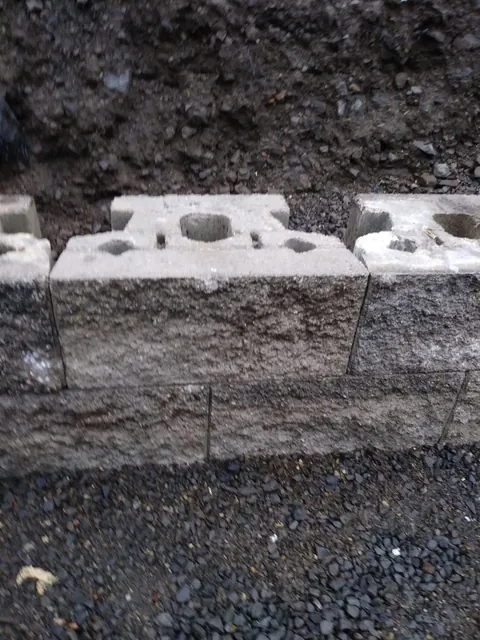 Textured landscape blocks form a low course against exposed soil, with the hollow block tops still visible.